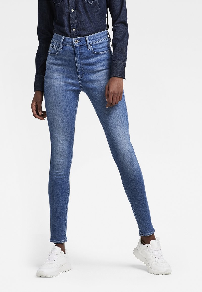 Jeans ajustados en denim azul claro, con una cintura alta, textura ligeramente desgastada y corte a la altura del tobillo, combinados con zapatillas blancas.