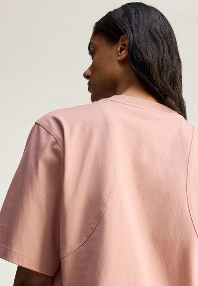 Young person with long dark hair wearing a loose light pink short-sleeve shirt, facing sideways against a plain light background.