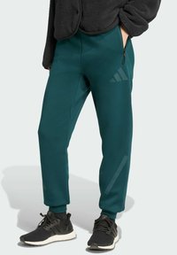 Zelené jogger nohavice vyrobené z hladkého materiálu, s bočnými vreckami na zips, rebrovými manžetami a nenápadným logom na dolnej časti nohavice. Skombinované s čiernymi teniskami.