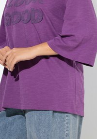 Camiseta púrpura de manga corta con un gráfico texturizado que dice "MIRA BIEN". Combinada con jeans azul de mezclilla, con un ajuste casual y holgado.