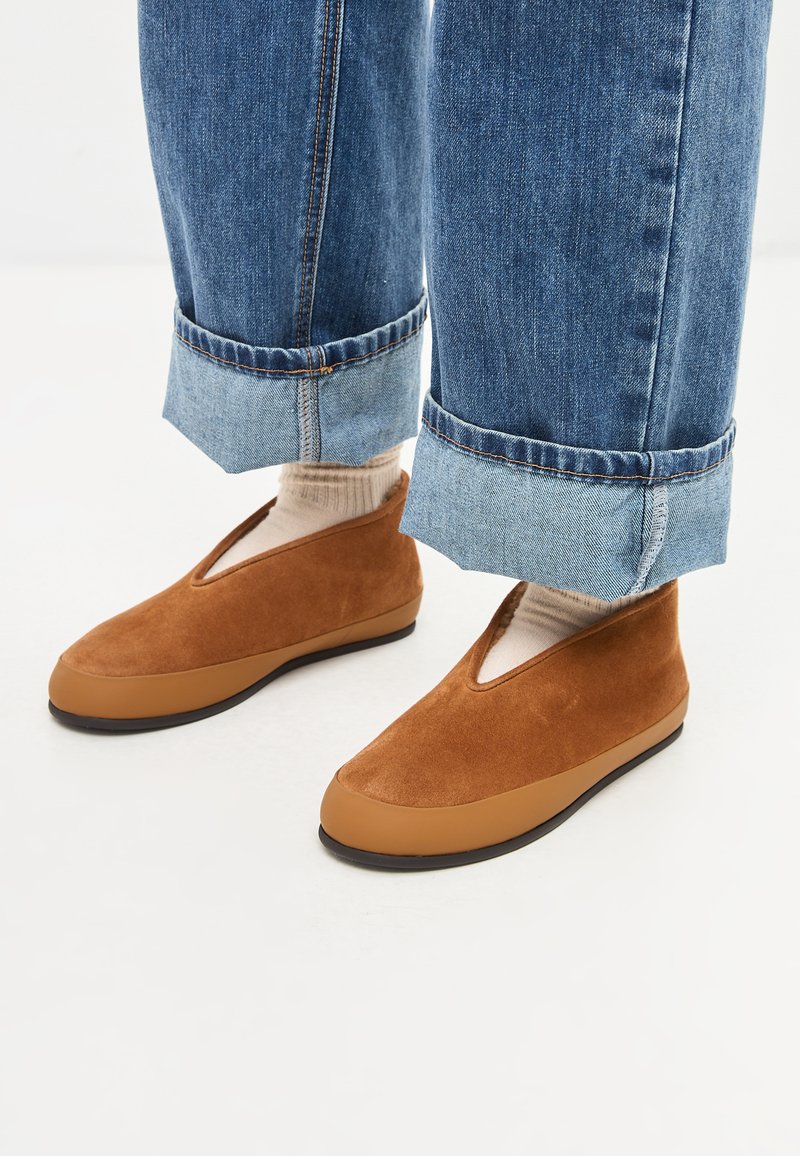 Bruine suède instap schoenen met een lichte V-vormige opening, op de achtergrond van opgerolde blauwe denimspijkerbroeken. Textuur bovenwerk, rubberen zool.