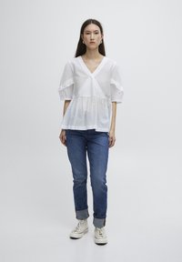 Haut peplum blanc à manches courtes avec un décolleté en V et des détails en volants. Porté avec un jean bleu et des baskets montantes blanches.