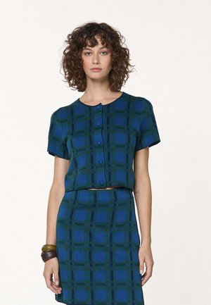 Femme aux cheveux bouclés portant un haut boutonné à manches courtes à carreaux bleu et vert, avec une jupe assortie et des bracelets marron à un poignet.