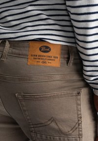 Jeans de mezclilla beige con parche de cuero marrón, detalles de costura visibles y dos bolsillos traseros. Camisa de rayas parcialmente visible encima.