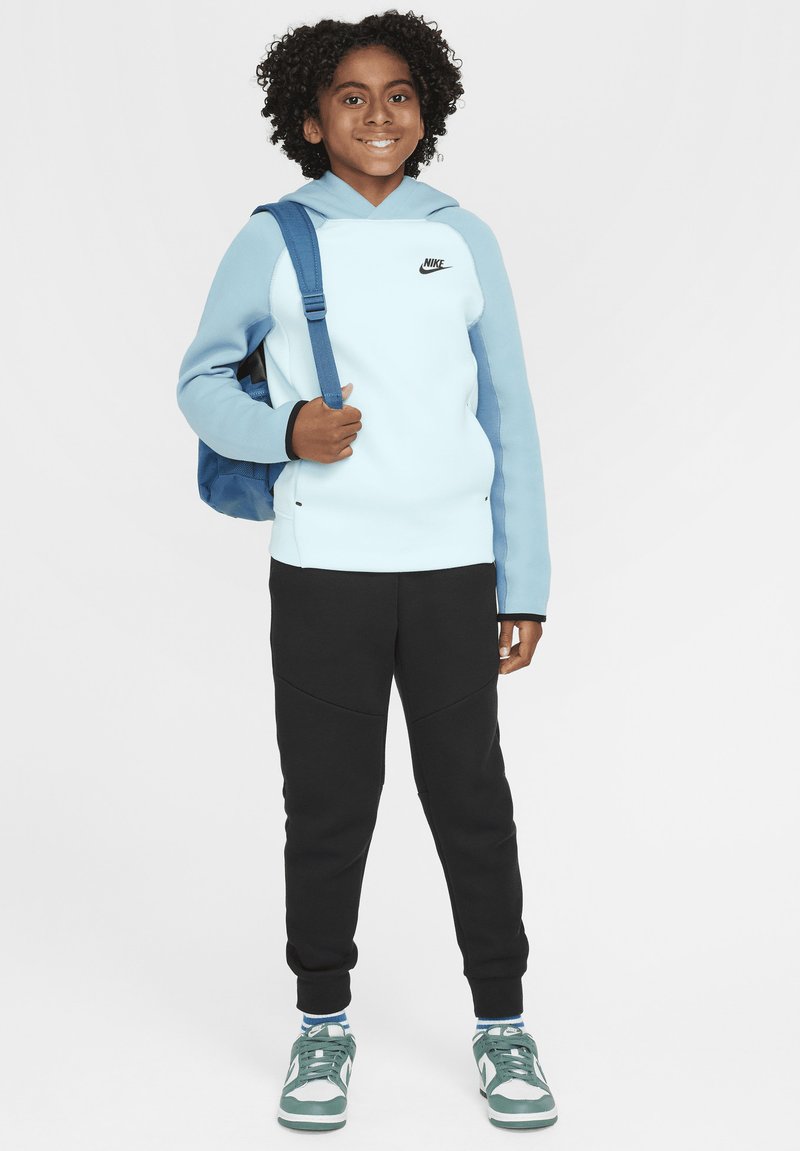 Sudadera con capucha de color azul claro con acentos negros, pantalones jogging negros y zapatillas verdes. Se lleva una mochila azul sobre un hombro.