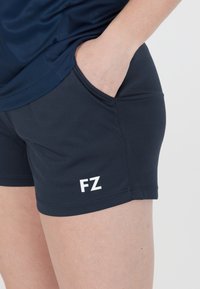 Person som bär marinfärgade sportiga shorts med "FZ"-logotyp, hand i fickan, och matchande marinblå topp mot en enkel vit bakgrund.