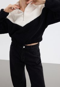 Chaqueta corta con cremallera en negro y crema, hecha de un material esponjoso, con un diseño en bloques de color, un cuello alto y costuras pronunciadas.