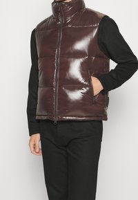 Brun, blank puffervest med høj krave, lynlås foran og sidelommer. Bæres over en sort langærmet skjorte og sorte bukser.