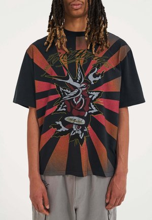 Osoba nosząca czarny, oversize'owy t-shirt z czerwonymi i pomarańczowymi promieniami oraz grafiką róży i ptaków na środku.