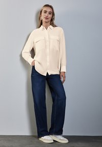 Camisa de botón de manga larga en color crema pálido con dos bolsillos frontales, combinada con jeans de pierna ancha en azul oscuro y zapatillas blancas.