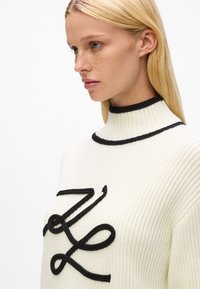 Pull en maille blanche épaisse avec col montant noir à côtes et lettres cursives noires brodées sur le devant. Tissu texturé, style ajusté.