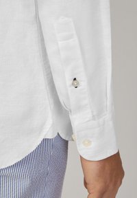 Manga de camisa de lino blanca con puño abotonado y botones claros, textura suave y dobladillo curvado. Pantalones a rayas azul y blanca visibles.