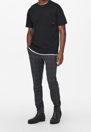 T-shirt nera a maniche corte con taschino sul petto e bordo collo bianco a contrasto, abbinata a pantaloni grigi slim-fit a quadri e stivaletti neri.