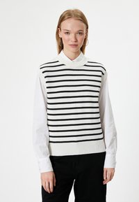 Jersey de punto sin mangas a rayas en blanco y negro, con un cuello alto y un dobladillo acanalado, capas sobre una camisa blanca de botones.