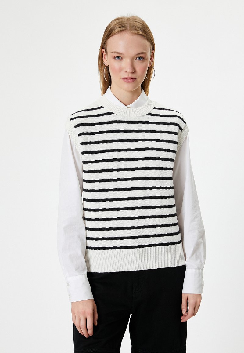 Jersey de punto sin mangas a rayas en blanco y negro, con un cuello alto y un dobladillo acanalado, capas sobre una camisa blanca de botones.
