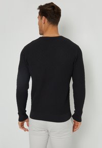 Un pull en tricot noir avec un motif en diamants texturé, un col rond, et des poignets et un ourlet côtelés, associé à un pantalon de couleur claire.