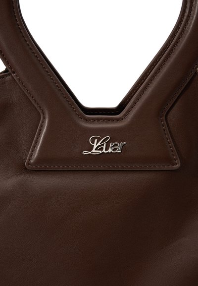 Borsa a mano in pelle marrone con un design strutturato, dotata di un manico intagliato e di un accento in logo argentato cucito sopra. Texture liscia.