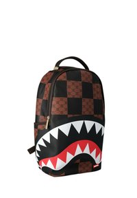 Mochila con patrón de cuadros marrón y negro con signos de dólar y un gran gráfico de boca de tiburón con dientes blancos y encías rojas.
