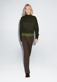 Dunkelgrüner Strickrollkragenpullover, übereinander getragen mit einem hellgrünen Top, kombiniert mit braunen Schlaghosen und dunklen Schuhen. Einfaches, strukturiertes Design.