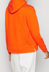 Oranje hoodie van zachte stof, met een capuchon met trekkoord, zijsplitten bij de zoom en geribbelde manchetten. Witte broek gedeeltelijk zichtbaar.