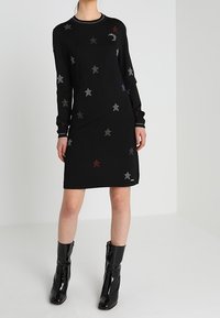 Abito nero a maglia con decorazioni di stelle multicolori, maniche lunghe e scollatura rotonda. Abbinato a stivali neri lucidi alla caviglia.