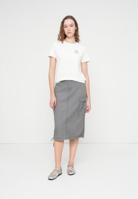 Λευκό βαμβακερό μπλουζάκι με μικρό κόκκινο λογότυπο, συνδυασμένο με γκρι φούστα μέχρι το γόνατο με τσέπη και πλευρικές σκισμές, φορεμένο με ασημένια φλατ παπούτσια.