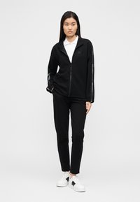 Zwarte fleece jas met ritssluiting en bijpassende broek, met een hoge kraag, ritssluiting aan de voorkant en omgeslagen mouwen. Witte accenten op de mouwen en schoenen.