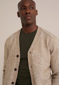 Béžový texturovaný kardigan s véčkovým výstřihem, žebrovanými manžetami a pěti tmavými knoflíky. Nositý přes tmavě zelené tričko na neutrálním pozadí.