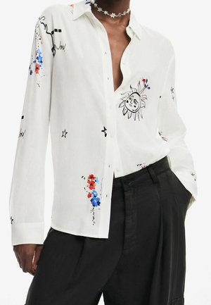 Hvid skjorte med lange ærmer og knapper, dekoreret med illustrationer af sol og stjerner samt farverige blomsterdetaljer, kombineret med sorte bukser med høj talje.