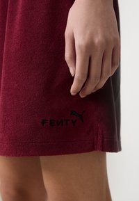Detailný záber ruky, ktorá drží okraj burgundského odevu, na ktorom je vyšitý nápis "FENTY" a logo Puma blízko spodného okraja.