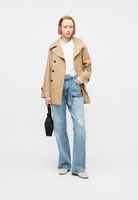 Γυναίκα με κόκκινα μαλλιά, που φοράει μπεζ καμπαρντίνα, λευκό πουκάμισο, ανοιχτό μπλε τζιν με σκισίματα και ζώνη, λευκά παπούτσια, κρατώντας μια μικρή μαύρη τσάντα.