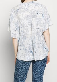 Top leggero e fluido realizzato in tessuto bianco con motivi floreali blu a spirale, maniche corte in stile mantella e scollatura arricciata.
