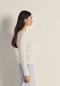 Pruunide pikkade juustega naine, kes kannab valget pikakäist toppi ja helehallid pükse, seisab külje pealt tavalise beežika tausta ees.