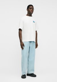 Άνδρας με γυαλιά, λευκό T-shirt με μικρό μπλε σχέδιο, ανοιχτό μπλε τζιν παντελόνι φαρδύ και μαύρα παπούτσια, στέκεται μπροστά σε απλό φόντο.