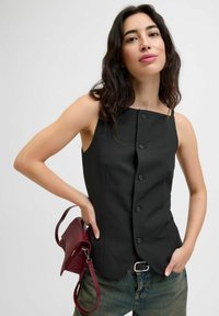 Kvinna med långt mörkt hår som bär en knappad svart ärmlös topp, blå jeans och håller en liten burgunderväskan bakom ryggen.