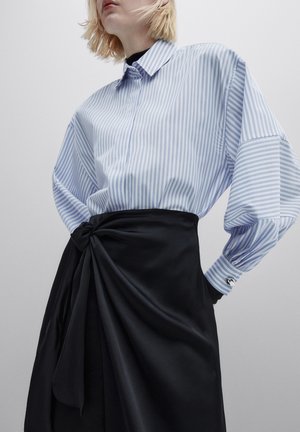 Femme portant une chemise à rayures bleues et blanches boutonnée, rentrée dans une jupe noire taille haute nouée à la taille.