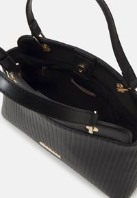 Sac à main noir avec texture côtelée verticale, ferrures dorées et deux poignées. L'intérieur comprend une poche zippée et une étiquette logo.