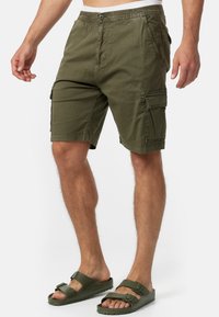 Hombre vestido con pantalones cortos de cargo verde olivo y sandalias a juego, de pie frente a un fondo blanco.
