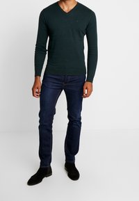 Maglione verde scuro a maniche lunghe con scollatura a V abbinato a jeans blu scuro e stivali in suede neri. Il maglione ha un design aderente e orlo a coste.