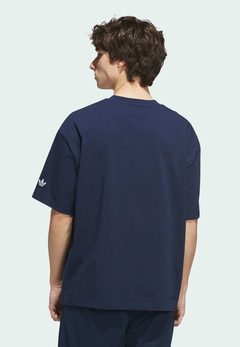 Jeune homme aux cheveux courts et bouclés portant un t-shirt bleu marine uni et un short foncé, vu de dos sur un fond clair.