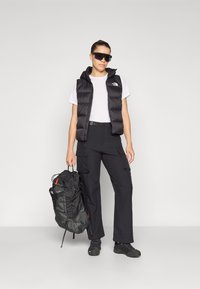 Zwarte puffer vest over een wit t-shirt, gecombineerd met zwarte cargo pants. De figuur houdt een zwart en grijze rugzak vast en draagt grote zonnebril.