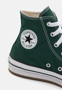 Zapatilla alta de lona verde con cordones blancos, logo de estrella negra, detalles de costura y suela de goma con rayas blancas y marrones.