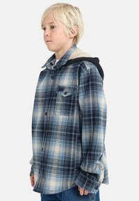 Camicia in flanella a quadri blu e crema con cappuccio nero. Presenta una chiusura a bottoni sul davanti e una tasca sul petto. Tessuto morbido e strutturato.