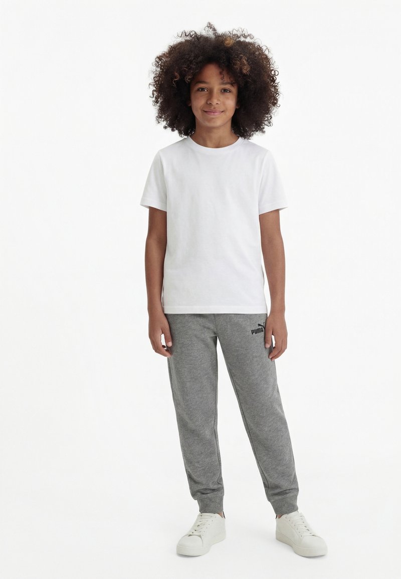 Vaikas su garbanotais plaukais, vilintis baltą marškinėlį, pilkas Puma sportines kelnes ir baltus sportinius batelius, stovintis priešais paprastą baltą foną.