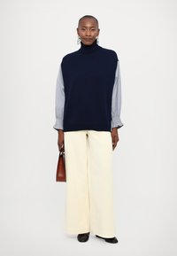 Navy knitted sleeveless turtleneck, paired with a light blue striped shirt, white wide-leg trousers, and a brown handbag. Black shoes.