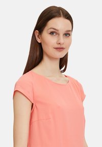 Korallenrosa Bluse mit kurzen Ärmeln, rundem Ausschnitt und dezentem Seitentaschen-Detail. Glatter Stoff mit leichtem Griff.