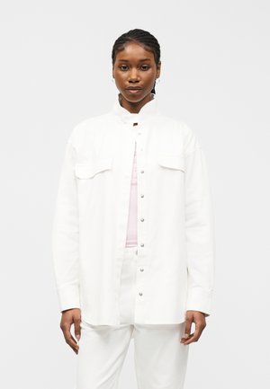 Γυναίκα με πλεξούδες, φοράει λευκό πουκάμισο με κουμπιά πάνω από ανοιχτό ροζ τοπ και λευκό παντελόνι, στέκεται μπροστά σε απλό λευκό φόντο.