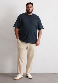 Donkerblauw T-shirt met korte mouwen en een borstzak, gecombineerd met beige broeken en witte sneakers met een tan-zool, tegen een effen achtergrond.