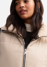 Woman with long dark hair wearing a beige quilted jacket with a leopard print lining and a white top underneath.