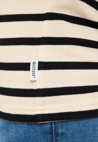 Čierno-biely pruhovaný rebrovaný sveter s priliehavým strihom. Má všitý štítok s nápisom "MUSTANG." Textil s textúrou a hladkým povrchom.